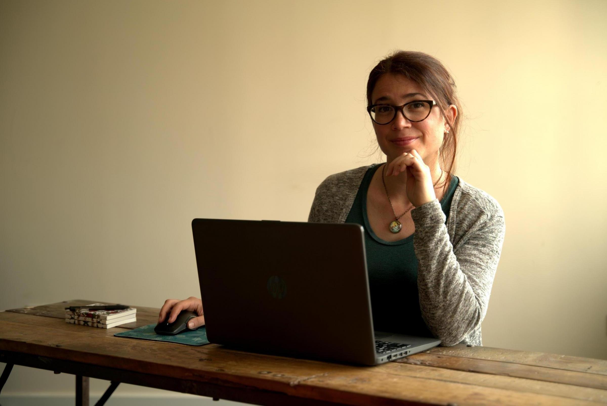 A picture of Alessandra sitting at a wooden desk, smiling at the camera and working at her laptop. She has brown hair and eyes and a fair complexion. She wears glasses and her hair is up.